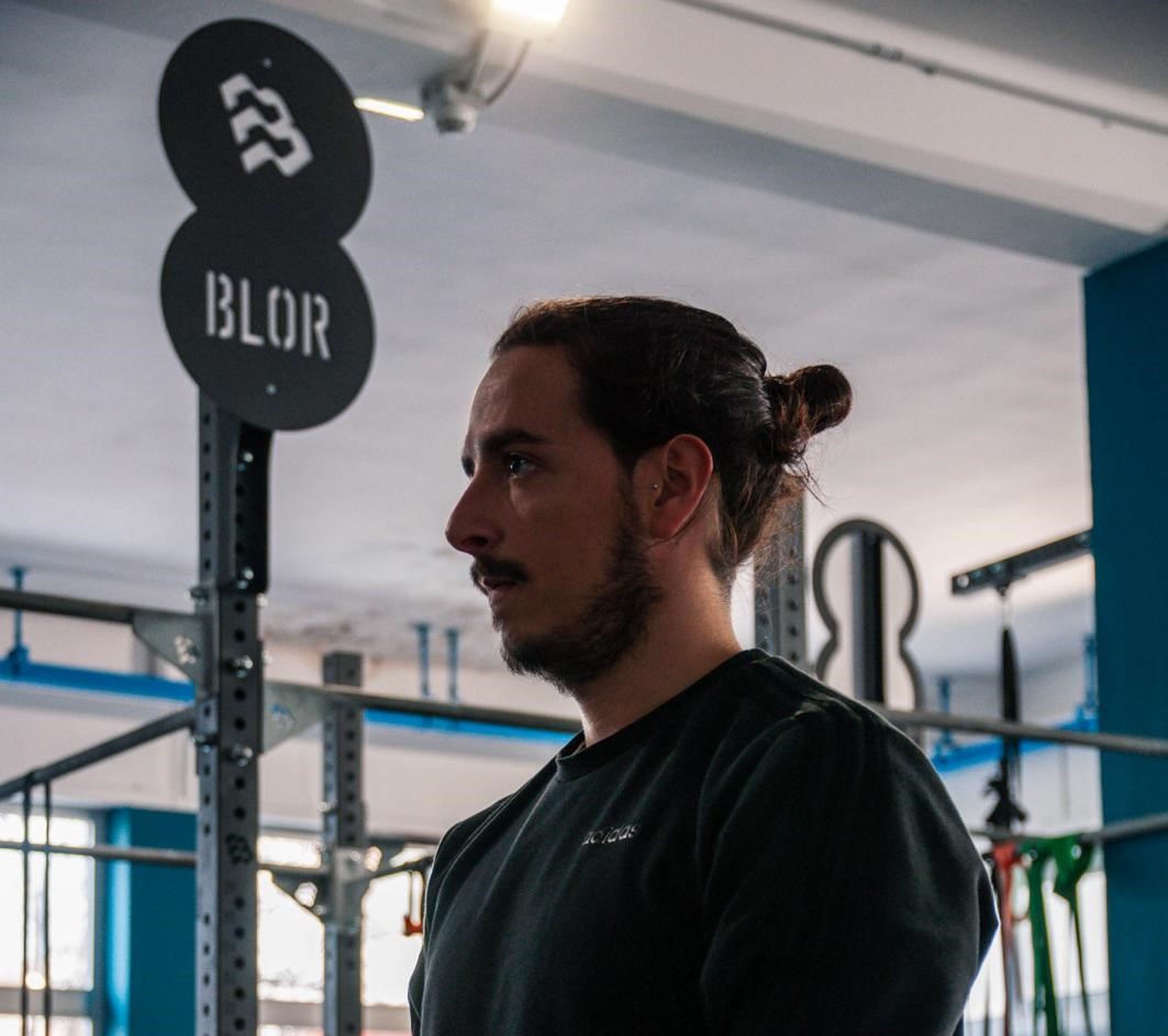 Un ragazzo col codino in ambiti da palestra, preso dal petto in su. Sullo sfondo si vedono attrezzi da palestra