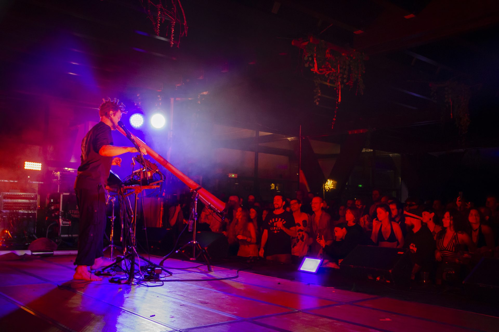 Una persona su un palco che suona un didgeridoo, sotto il pubblico in penombra 