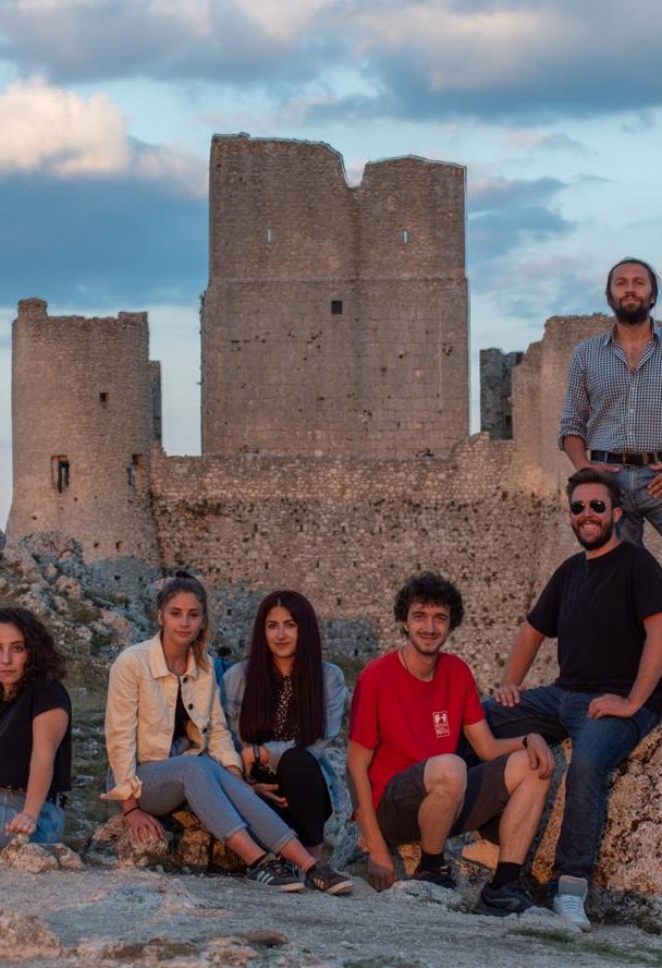 Gruppo di giovani in posa, alcuni seduti e altri in piedi, con una rocca sullo sfondo, al tramonto