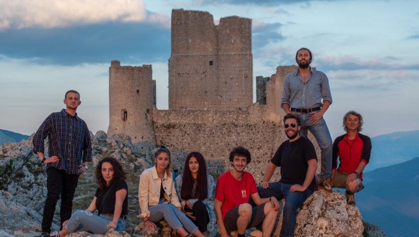 Gruppo di giovani in posa, alcuni seduti e altri in piedi, con una rocca sullo sfondo, al tramonto