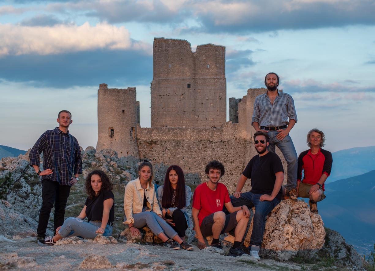 Gruppo di giovani in posa, alcuni seduti e altri in piedi, con una rocca sullo sfondo, al tramonto