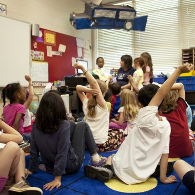 Dei bambini seduti a terra in una classe molto decorata, con una macchinina di carta che pende dal soffitto, l'insegnante è in fondo, circondata dagli alunni