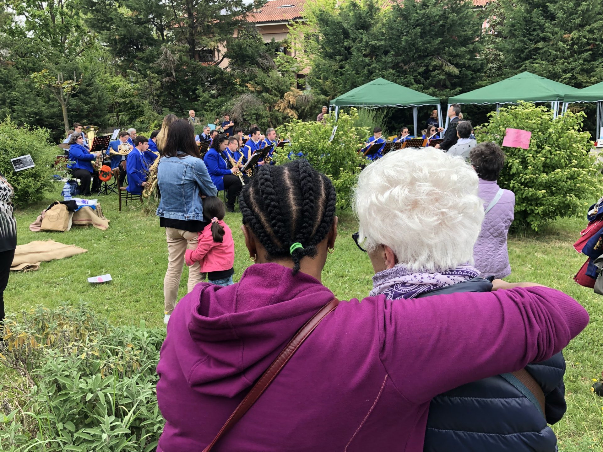 Due persone di spalle che si abbracciano, sullo sfondo persone in un prato, probabilmente a una fiera, con gazebi