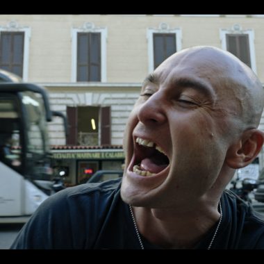 un primo piano di un uomo che urla, dietro a lui un palazzo