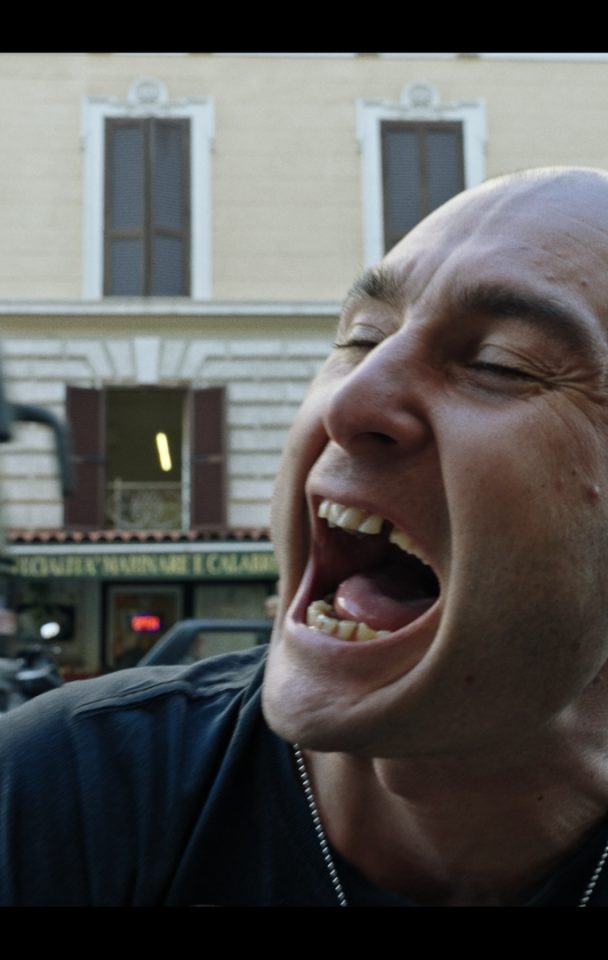 un primo piano di un uomo che urla, dietro a lui un palazzo