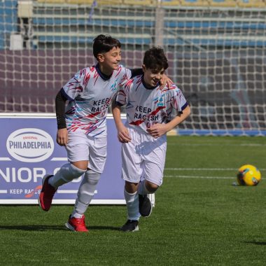 Due ragazzini in tenuta da calcio, sullo sfondo una rete e una palla, dietro si intravede un cartello con scritto philadelphia junior cup