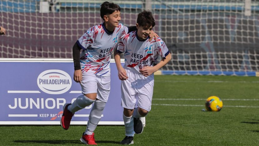 Due ragazzini in tenuta da calcio, sullo sfondo una rete e una palla, dietro si intravede un cartello con scritto philadelphia junior cup