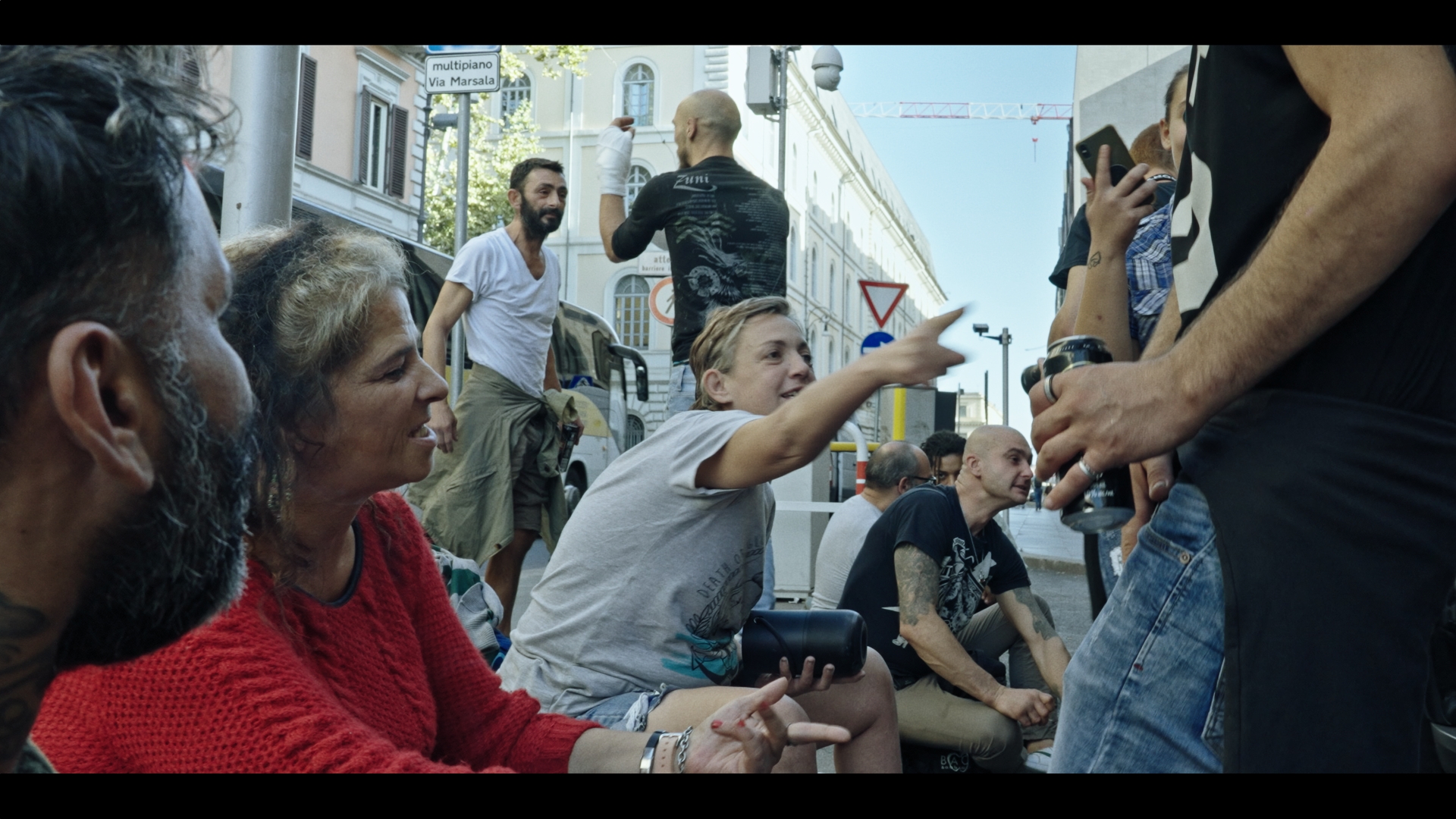 Delle persone sedute parlano, una indica con il dito, due sullo sfondo in piedi parlano