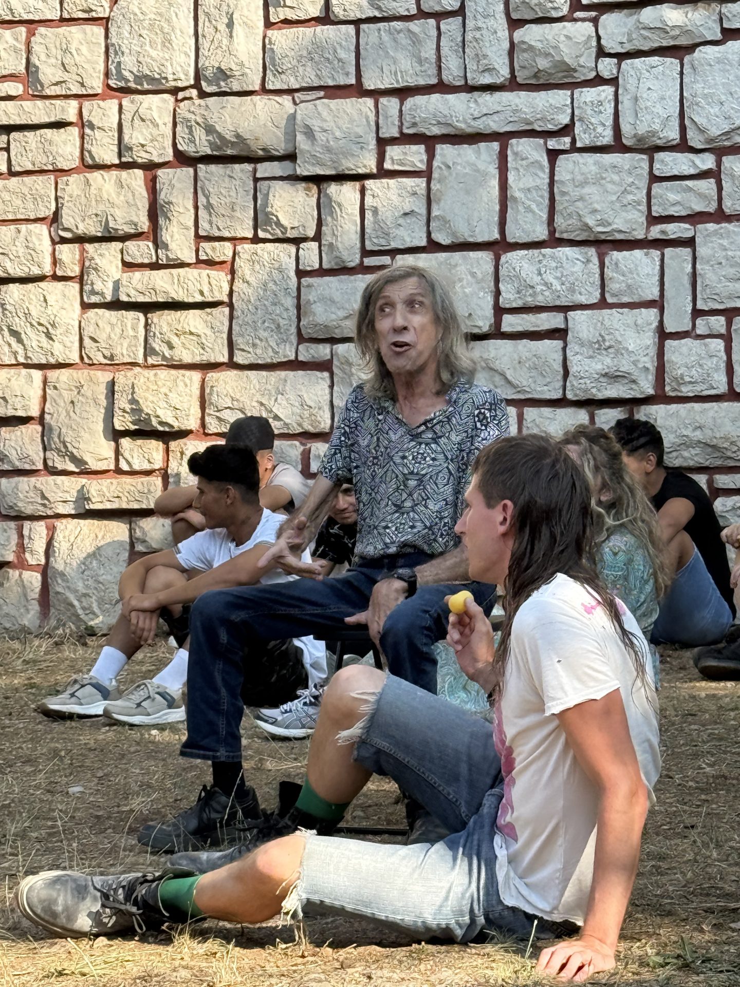 Una persona seduta su uno sgabello, con attorno altre persone sedute a terra, dietro un muro in pietra