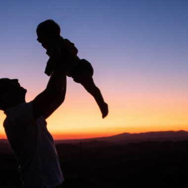In controluce, al tramonto, un padre che alza in alto il proprio figlio, guardandolo