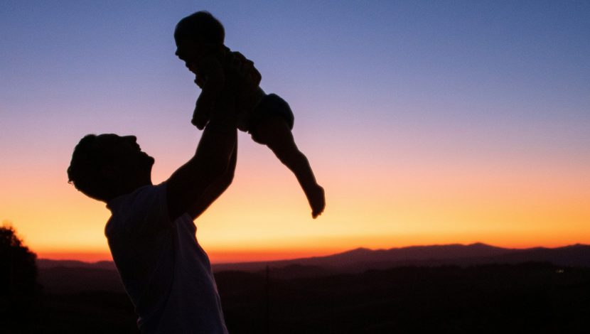 In controluce, al tramonto, un padre che alza in alto il proprio figlio, guardandolo