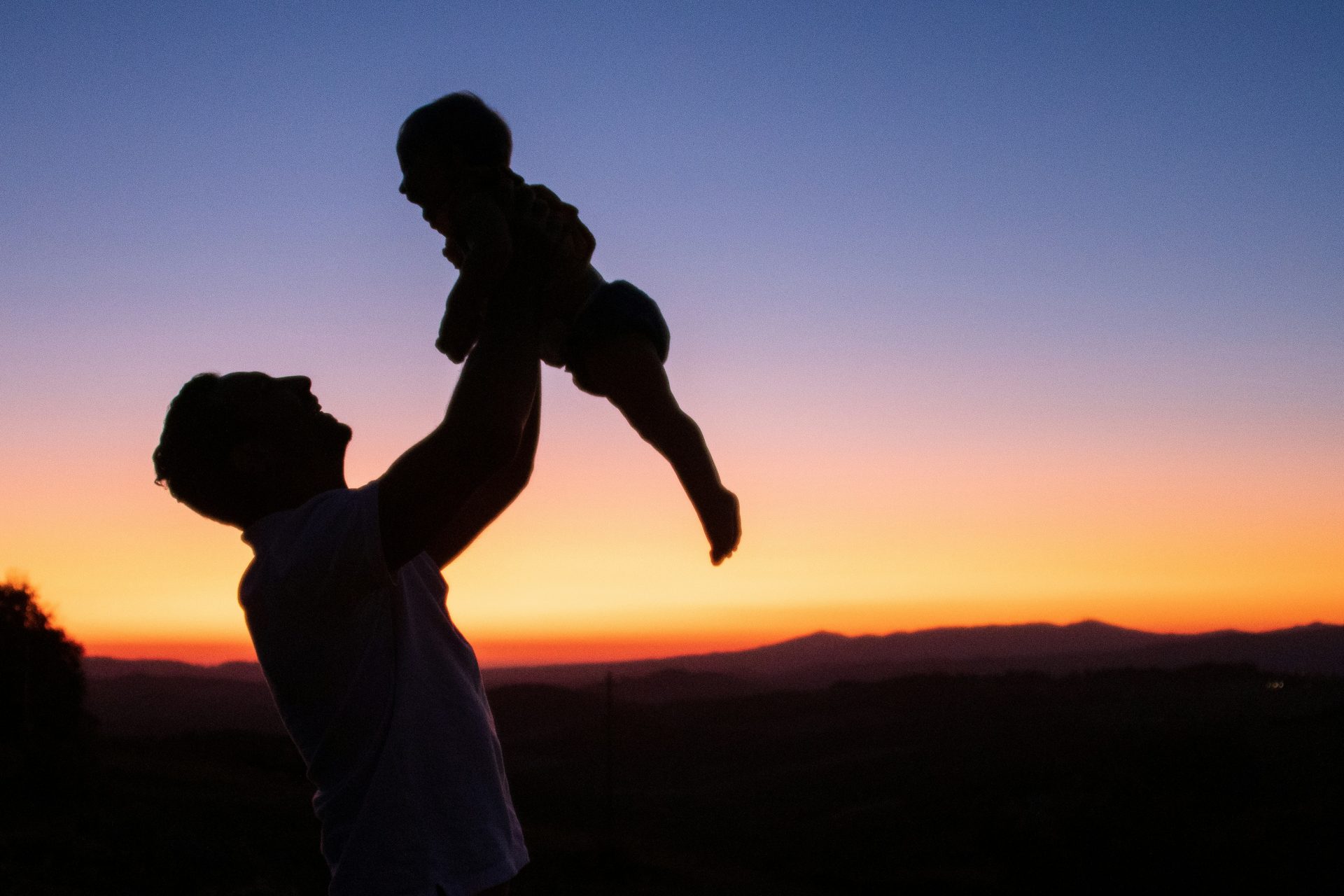 In controluce, al tramonto, un padre che alza in alto il proprio figlio, guardandolo