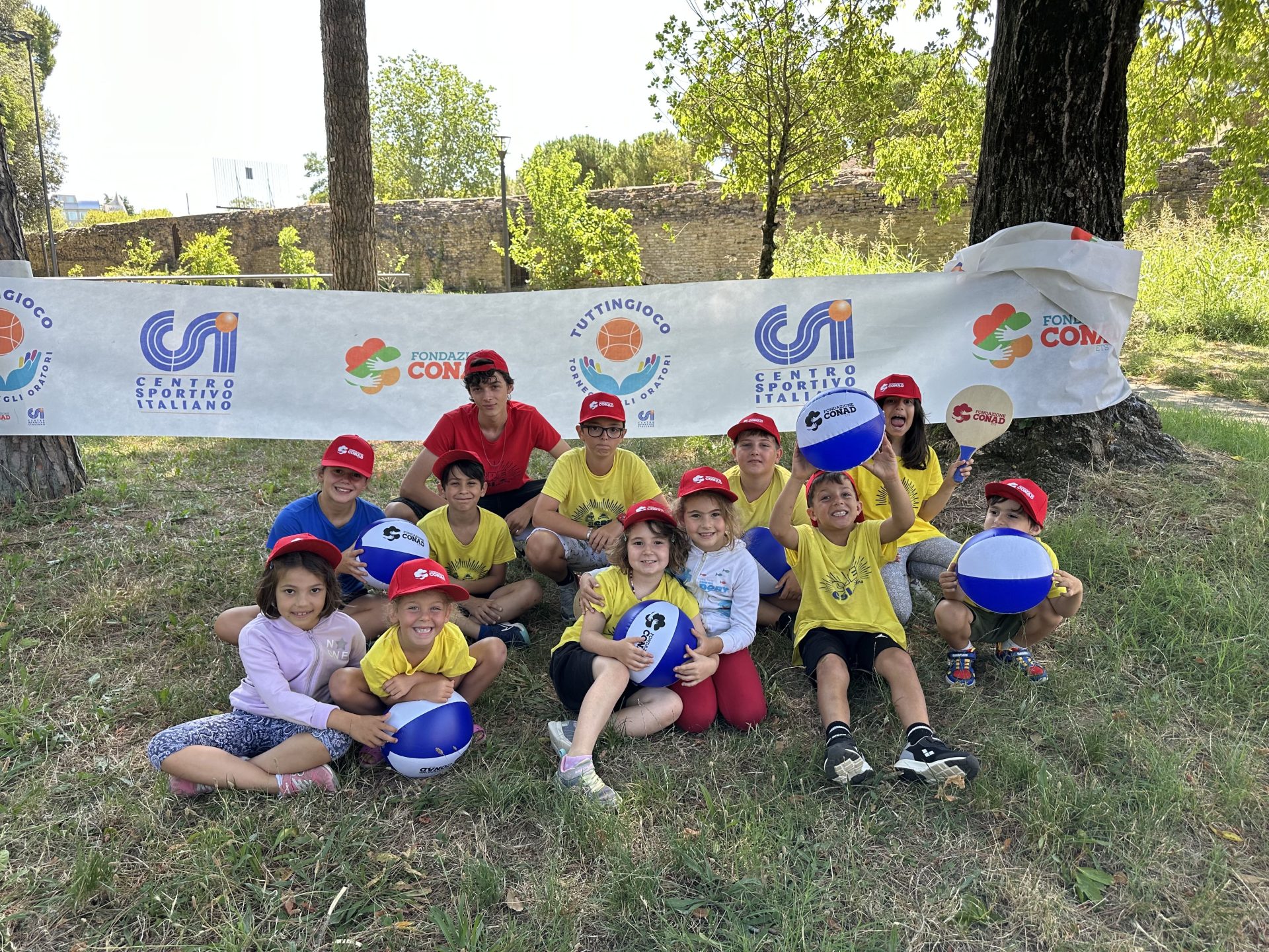 Alcuni bambini in gruppo seduti sull'erba, hanno delle palle in mano. Dietro uno striscione con scritto "csi"
