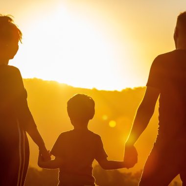 Una donna e un uomo tengono per mano un bambino che sta in mezzo, sono controluce, al tramonto