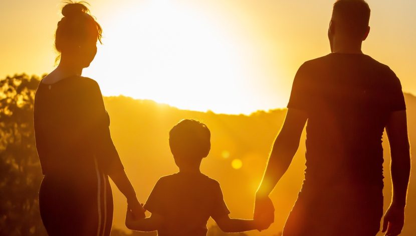 Una donna e un uomo tengono per mano un bambino che sta in mezzo, sono controluce, al tramonto