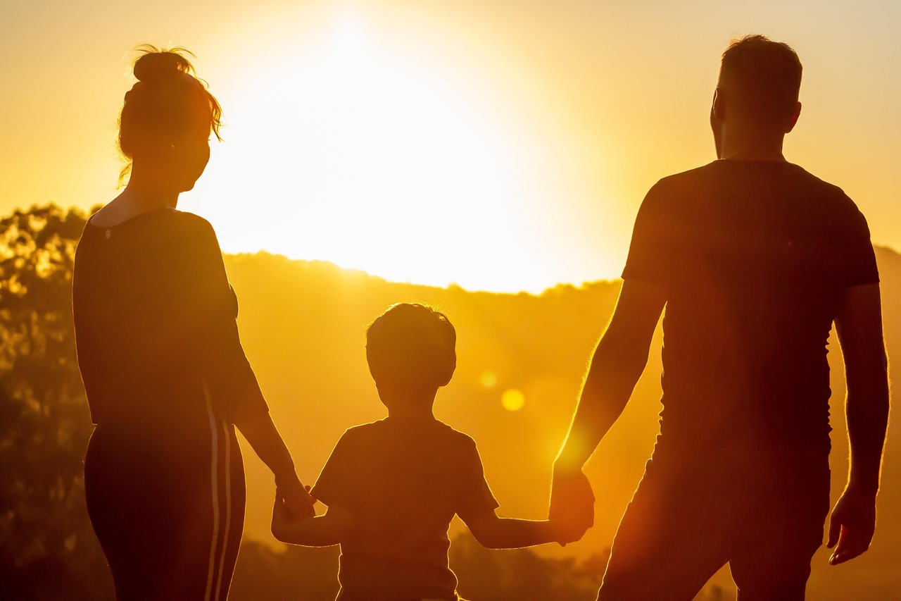 Una donna e un uomo tengono per mano un bambino che sta in mezzo, sono controluce, al tramonto